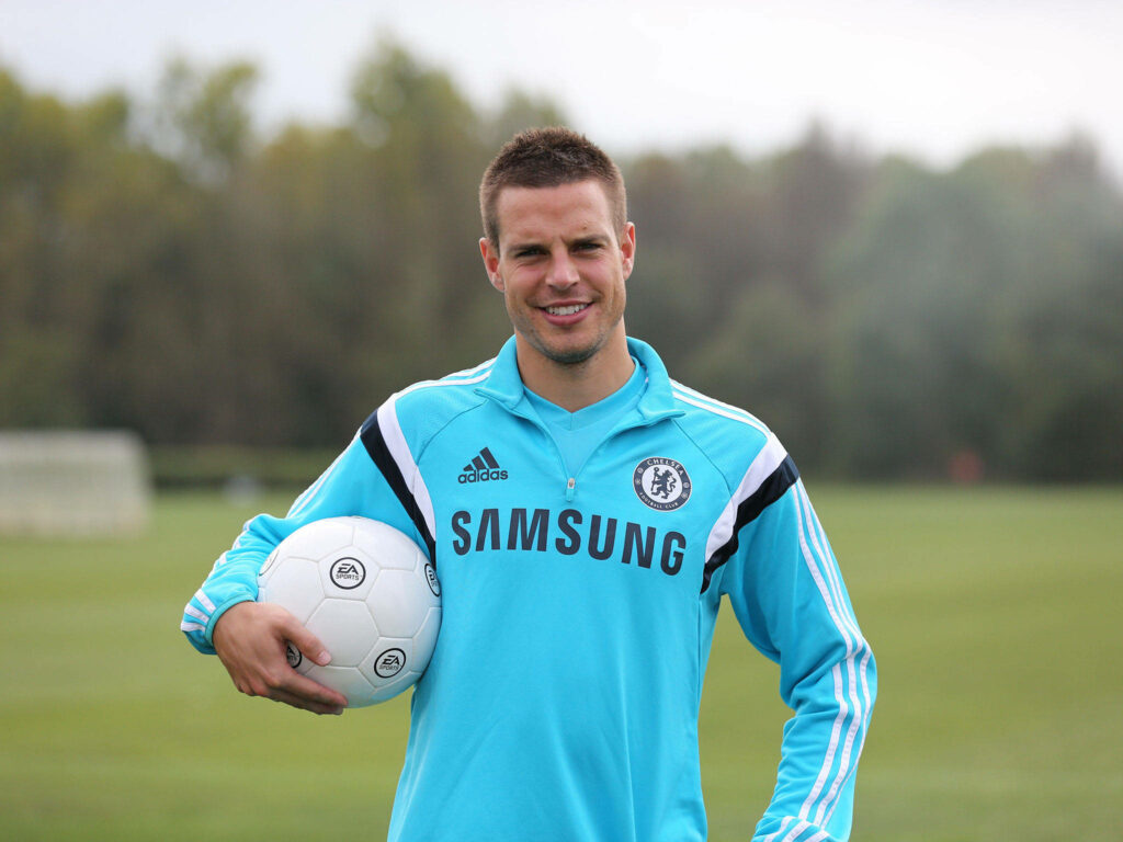 César Azpilicueta in light blue Chelsea jacket holding football on green field with trees background Wallpaper
