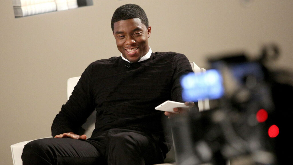 Chadwick Boseman smiling seated in black sweater modern minimalistic professional setting wallpaper