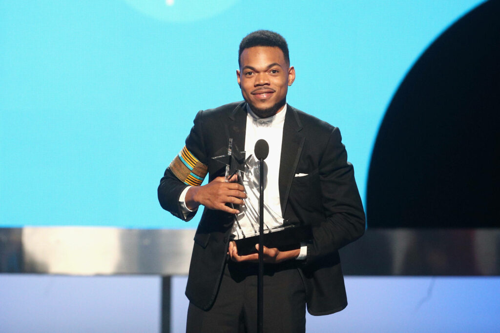 Chance The Rapper in black tuxedo holding award smiling on stage with vibrant blue background wallpaper