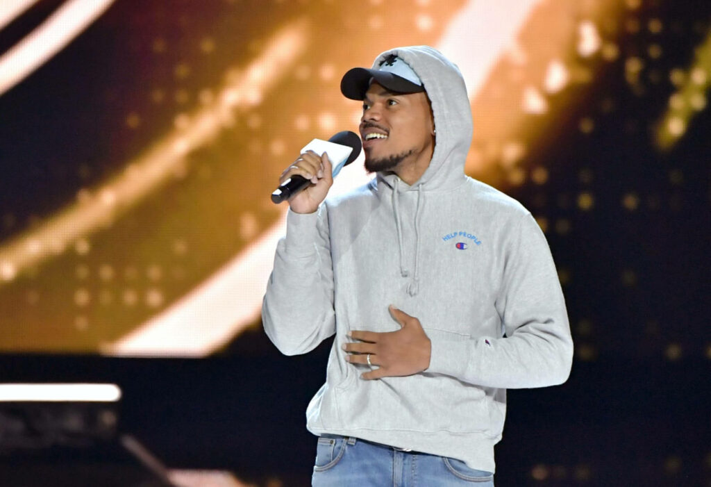 Chance The Rapper on stage wearing gray hoodie and cap speaking into microphone with gold background Wallpaper