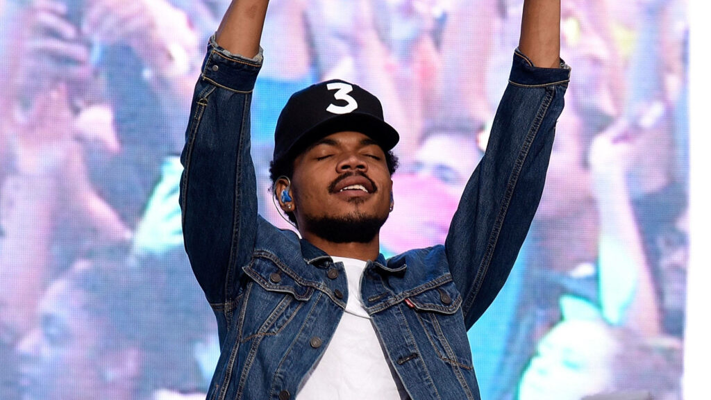 Chance The Rapper in black cap and denim jacket raising arms on stage with energetic crowd background Wallpaper