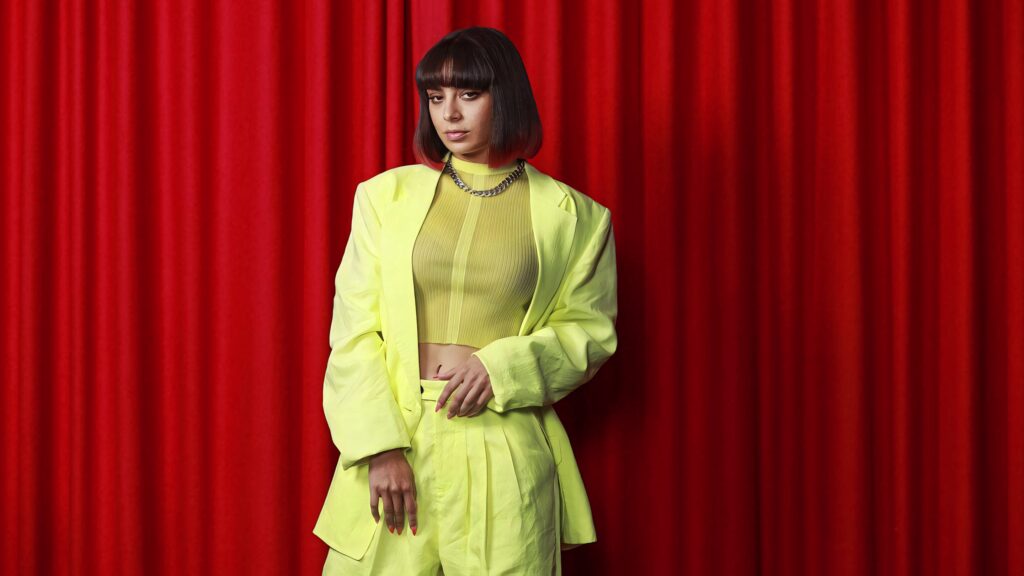 Charli XCX in neon yellow outfit standing confidently before red curtains in Sydney wallpaper background