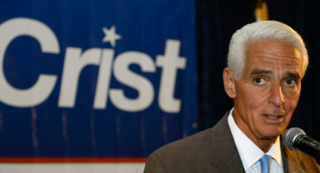Charlie Crist speaking at blue campaign backdrop in professional attire with focused expression Wallpaper