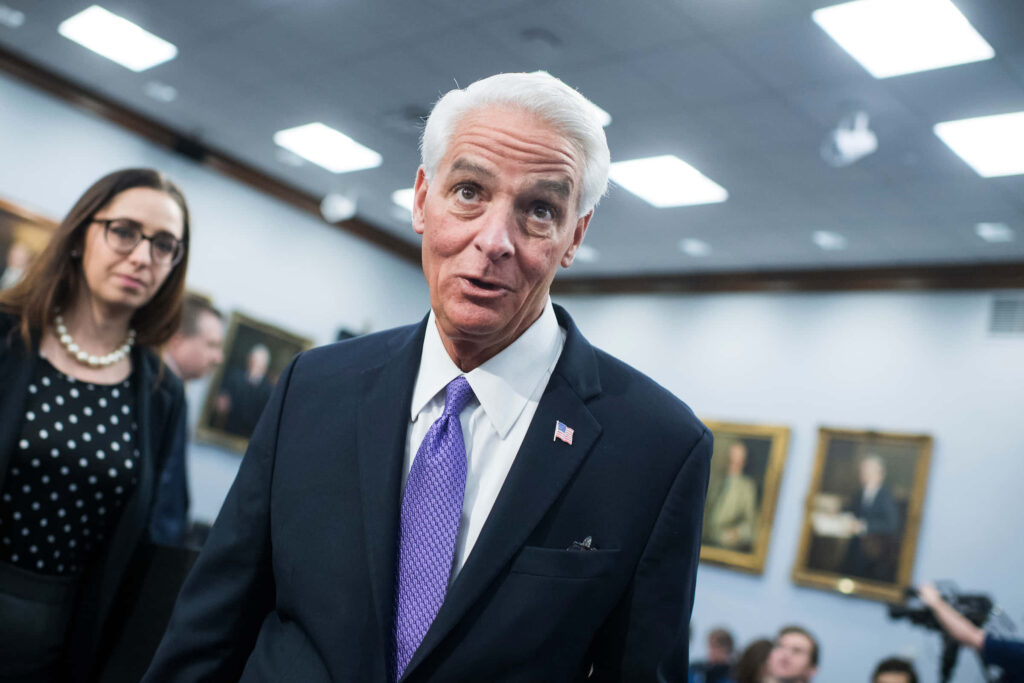 Charlie Crist in dark suit with purple tie and flag pin reflecting in formal setting with portraits Wallpaper