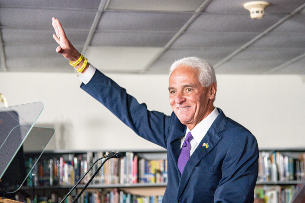 Charlie Crist smiling waving in blue suit purple tie with bookshelves bright library background wallpaper