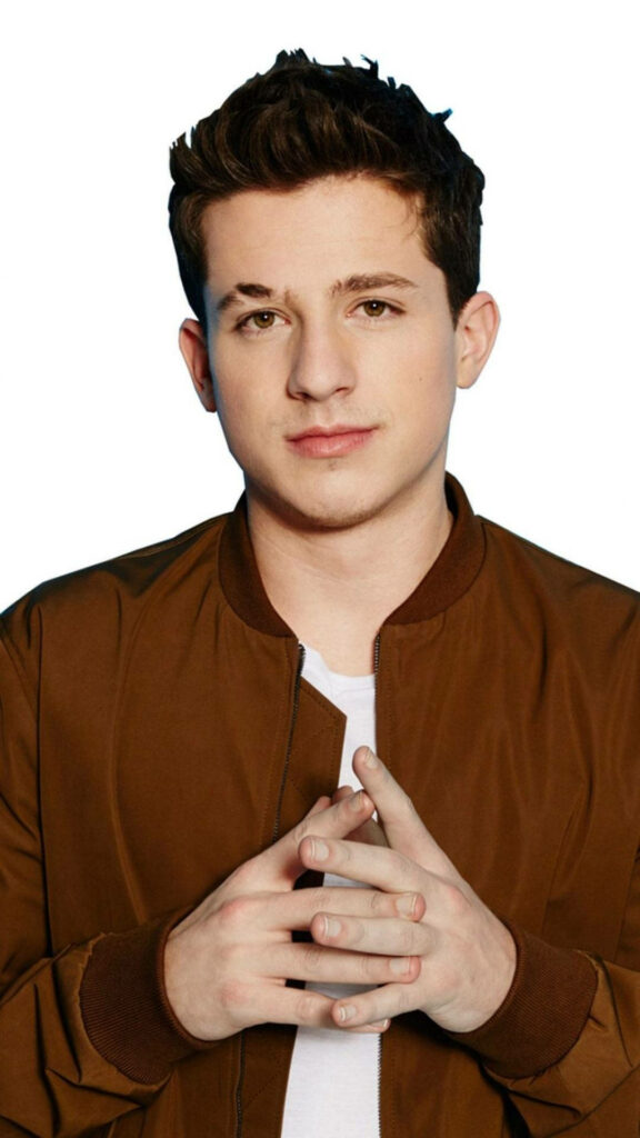 Charlie Puth in brown jacket with hands clasped against plain background stylish wallpaper