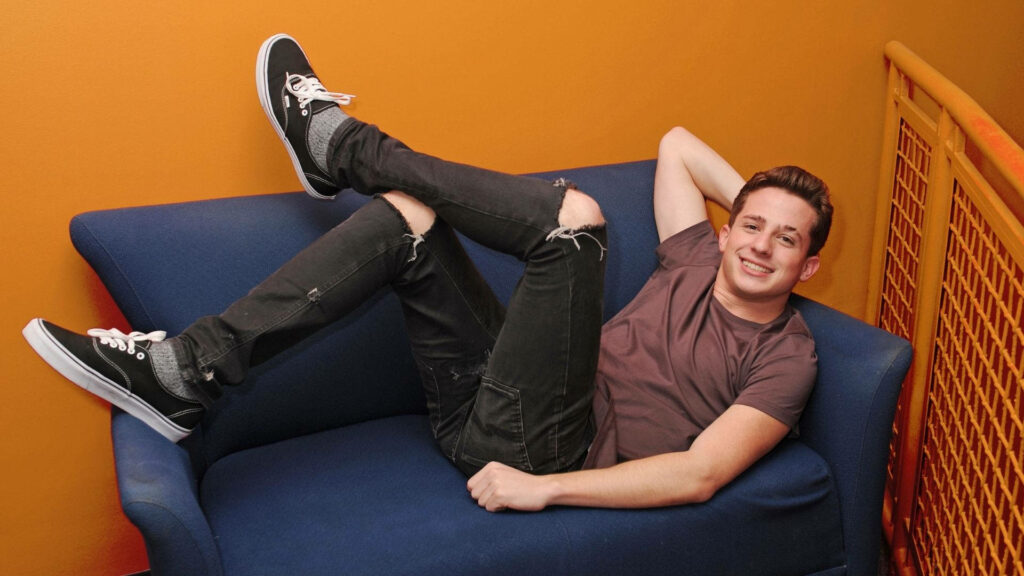 Charlie Puth relaxing smiling on blue sofa vibrant orange background cheerful wallpaper background