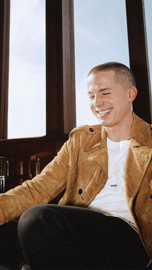 Charlie Puth smiling brightly in tan jacket and white shirt in warm cozy room wallpaper background
