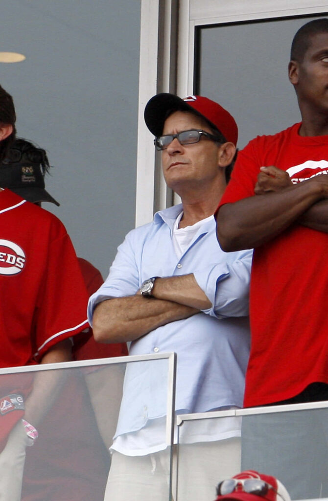 Charlie Sheen at baseball game wearing Cincinnati Reds cap and sunglasses with fans in red shirts Wallpaper