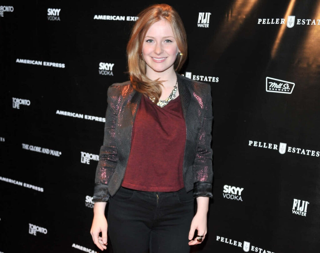 Charlotte Arnold in shimmering blazer and maroon top at branded event backdrop with confident pose Wallpaper