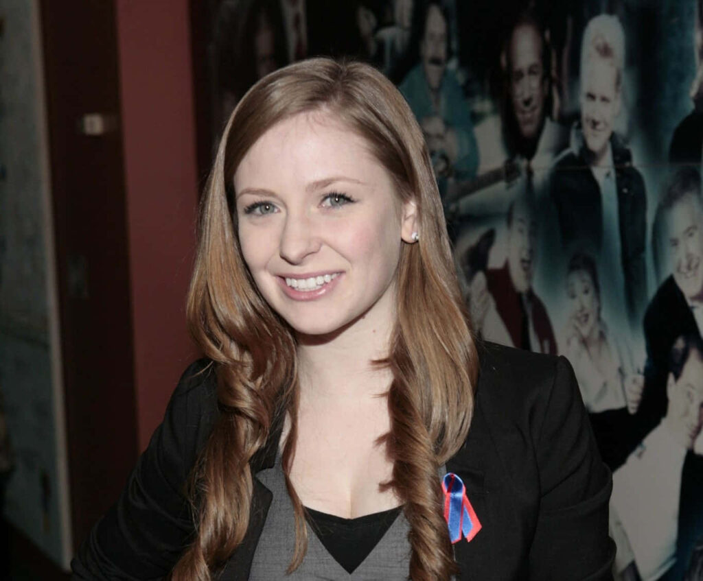 Charlotte Arnold smiling at event wearing ribbon on blazer with lively collage background wallpaper