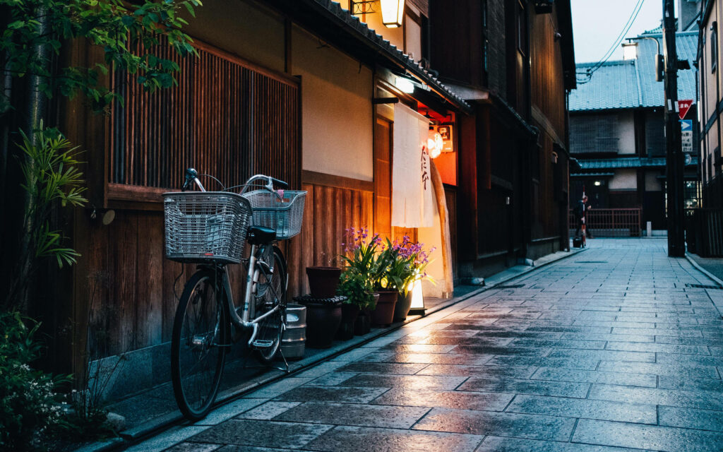 Bicycle by wooden building on cobblestone street with warm lantern light and flowers at dusk Wallpaper