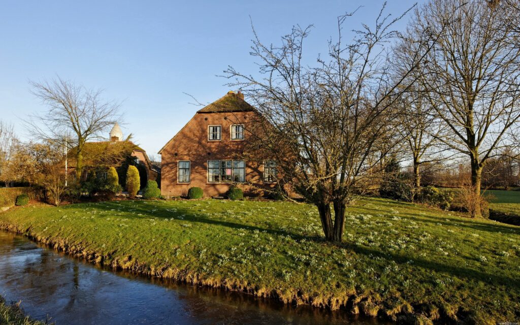 Charming brick house by narrow water channel in lush spring garden with wildflowers and trees Wallpaper