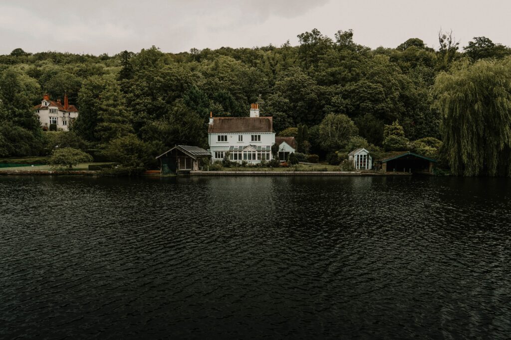 Charming lakeside house surrounded by lush trees calm water reflecting overcast sky peaceful scene Wallpaper