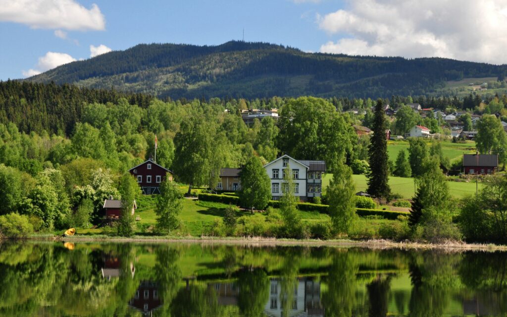 Charming lakeside houses with lush trees and majestic Alps reflected in calm water under blue sky Wallpaper