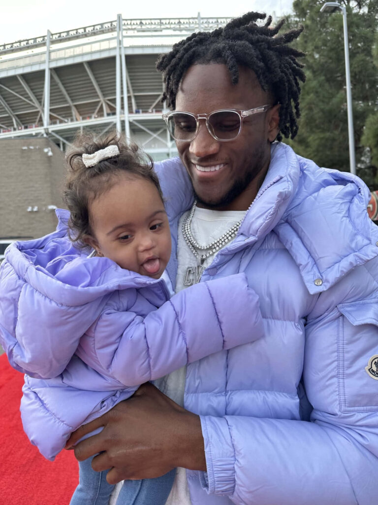 Charvarius Ward and daughter smiling in matching purple jackets outdoors near stadium wallpaper background