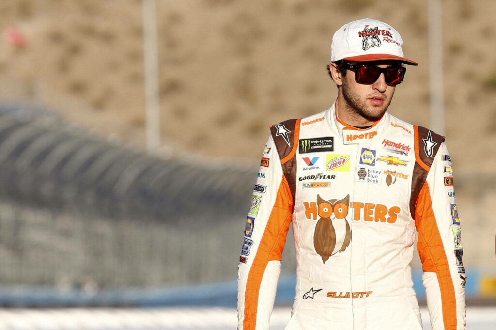 Chase Elliott in Hooters racing suit on racetrack with sponsor logos and determined expression Wallpaper