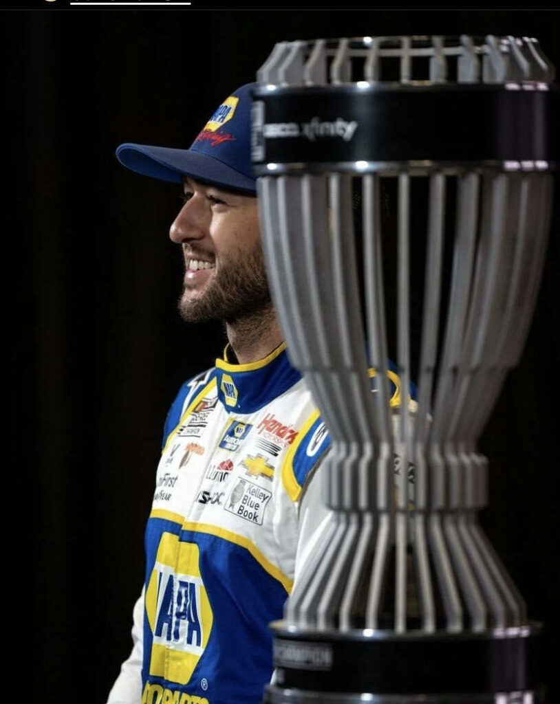Chase Elliott in blue yellow NAPA racing suit smiling beside detailed NASCAR trophy on dark background Wallpaper