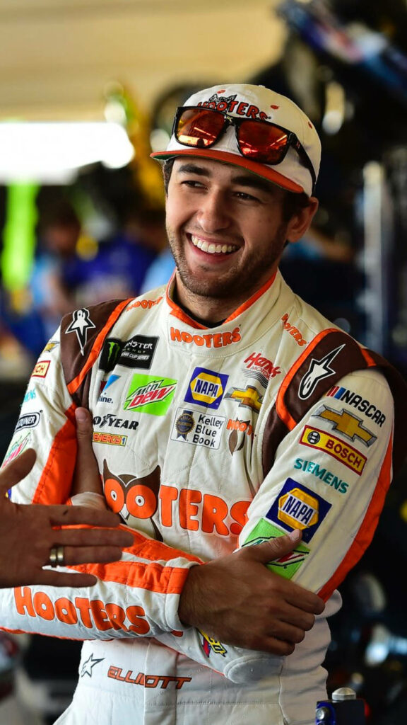 Chase Elliott smiling in white and orange Hooters racing suit in NASCAR pit area wallpaper background