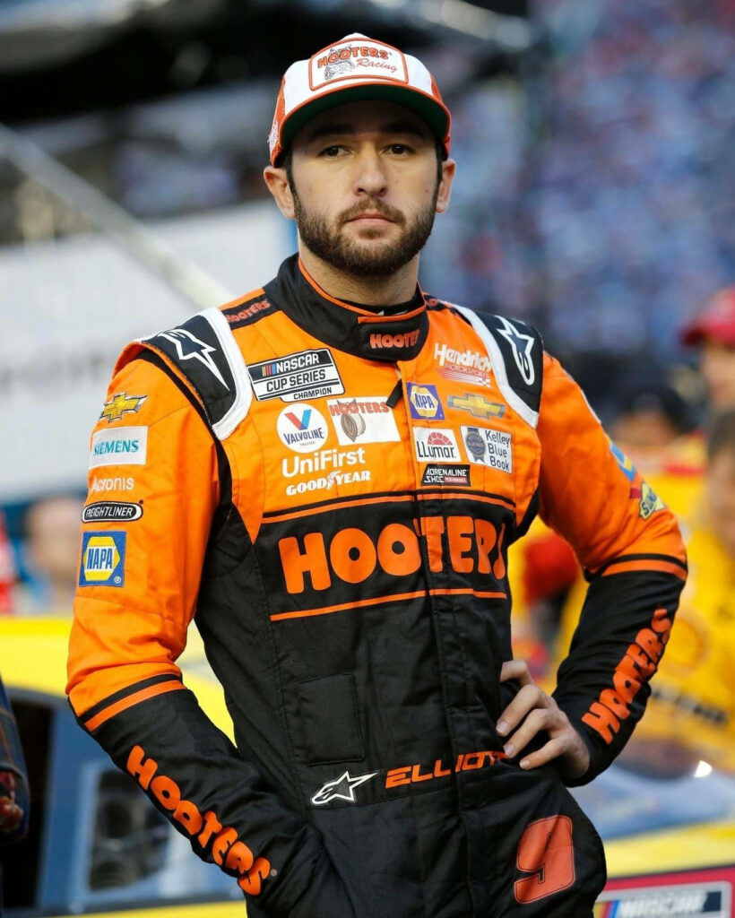 Chase Elliott in vibrant orange Hooters racing suit stands focused by his NASCAR race car wallpaper