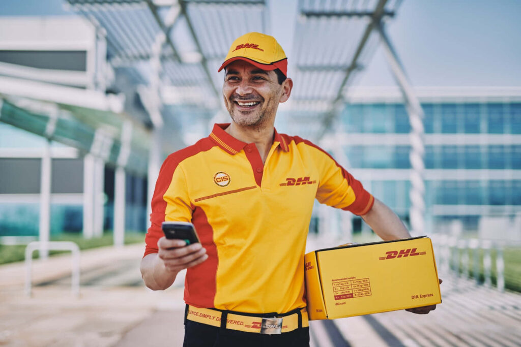 Cheerful DHL delivery man in uniform holding package and phone outside modern glass building Wallpaper