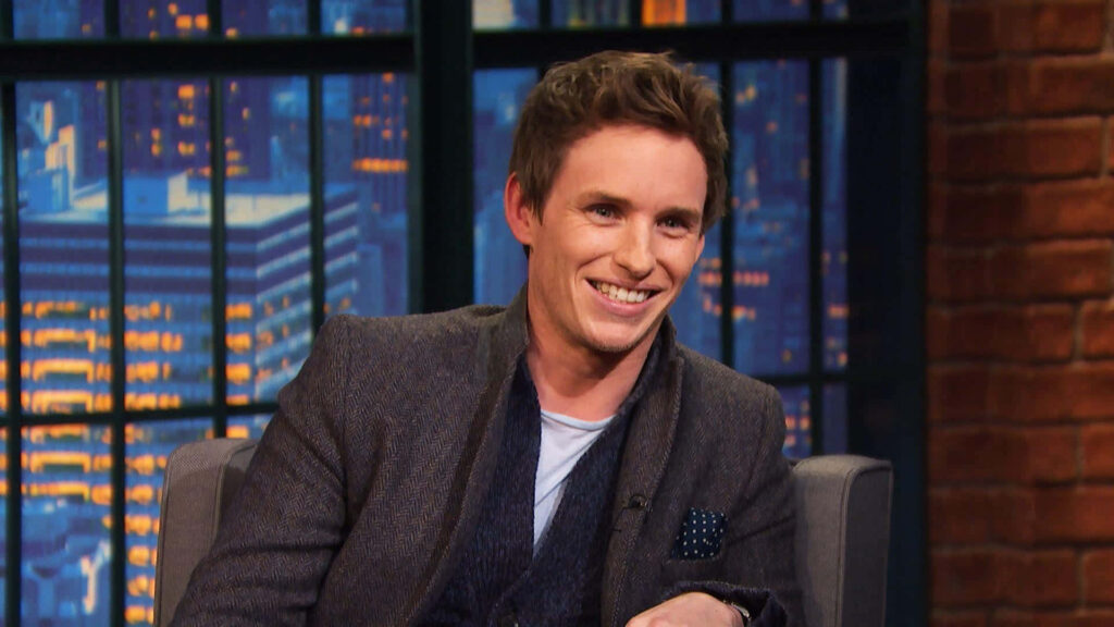Eddie Redmayne smiling during cozy urban TV interview with warm lighting and cityscape view Wallpaper