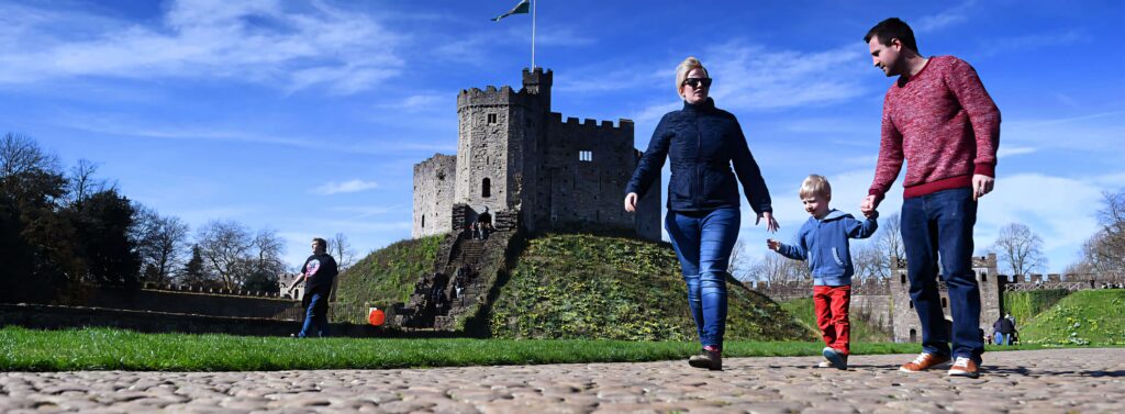 Cheerful family walking cobblestone path by historic Cardiff Castle under clear blue sky Wallpaper