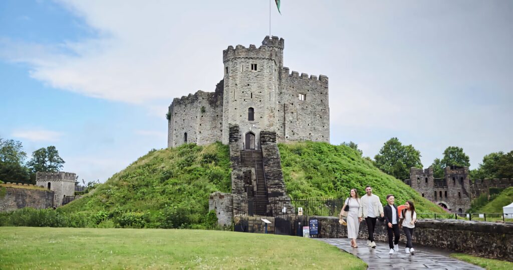 Cheerful family walking stone path toward historic Cardiff Castle with green landscape and sky Wallpaper