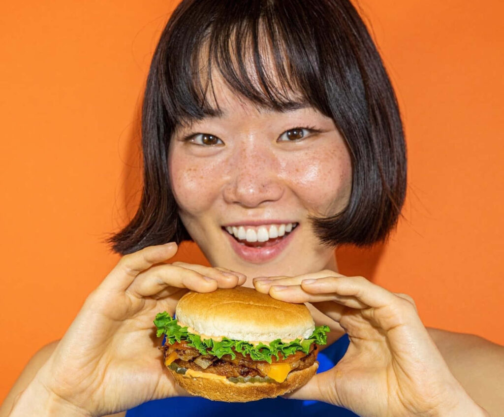 Cheerful Burger Lover with Fresh Toppings on Vibrant Orange Wallpaper