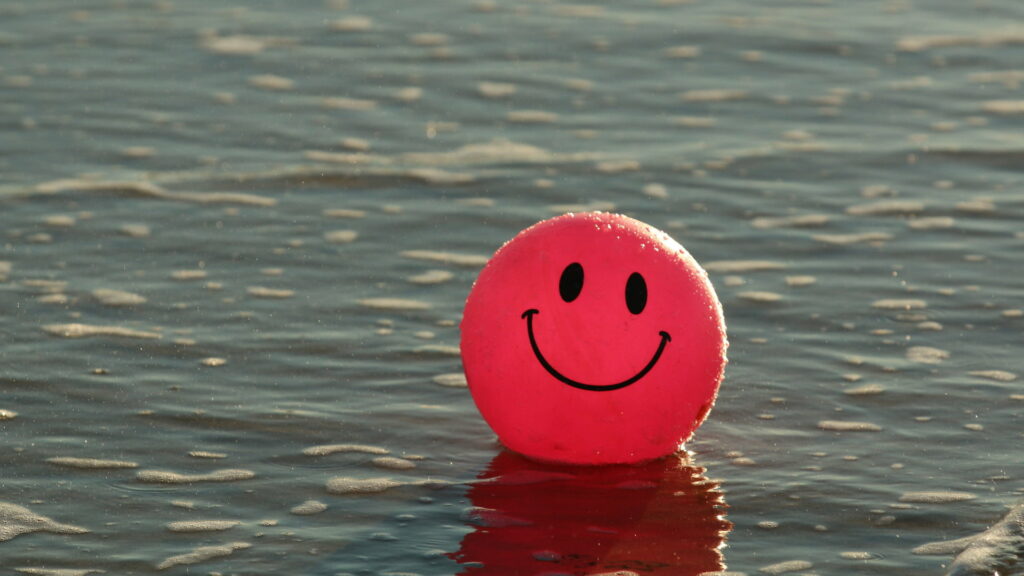Vibrant red smiley face ball floating on calm ocean water bright cheerful HD wallpaper background