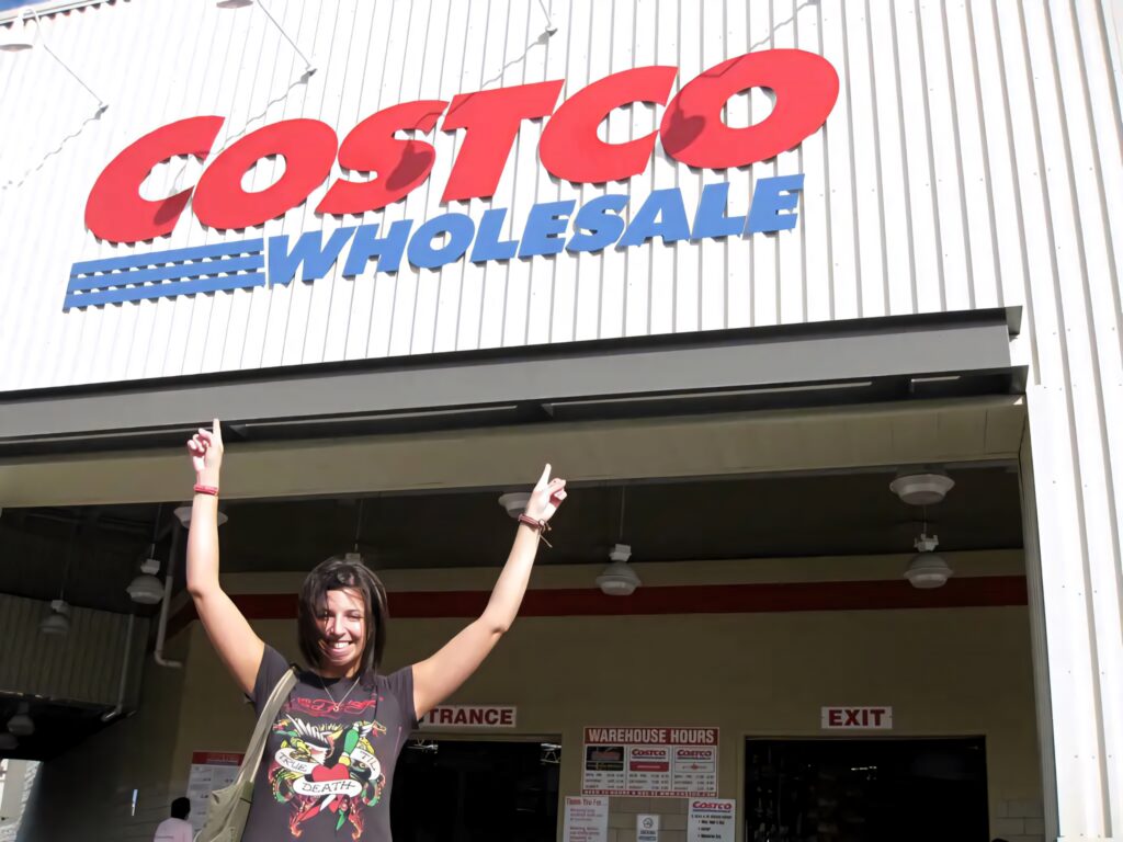 Cheerful woman pointing at large Costco Wholesale sign sunny day warehouse entrance background Wallpaper