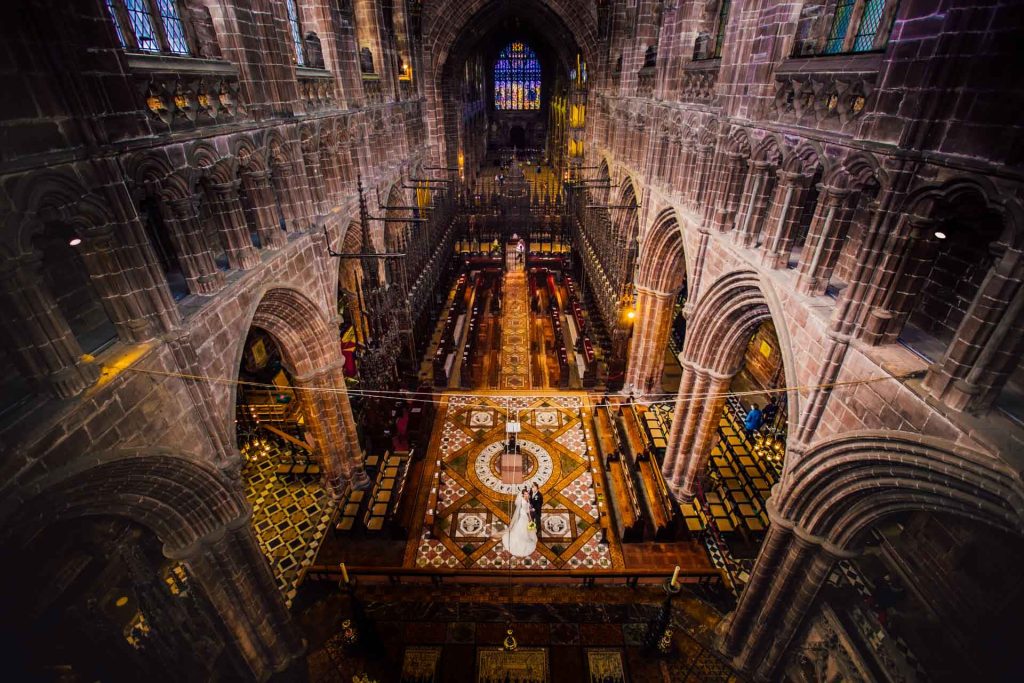 Bride and groom on intricate mosaic floor beneath Gothic red sandstone arches at Chester Cathedral interior Wallpaper