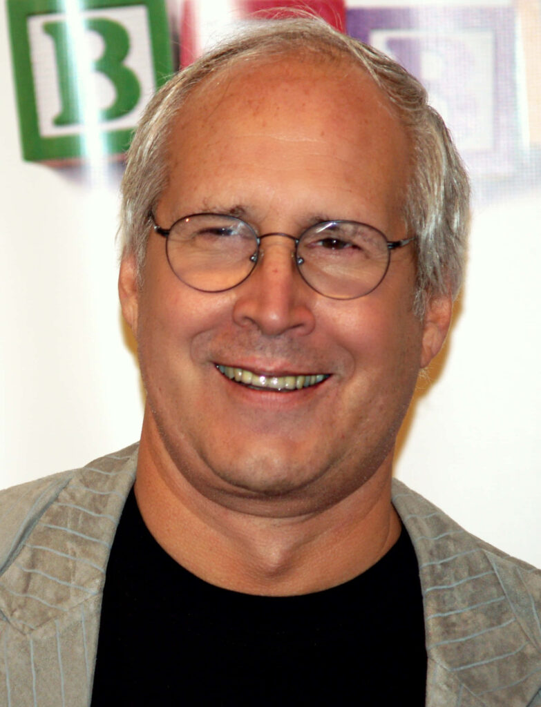 Chevy Chase smiling wearing glasses and light pinstriped jacket with colorful blurred background Wallpaper