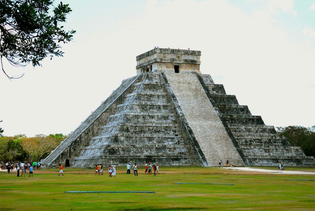Chichén Itzá Mayan pyramid with stone steps and visitors against pale sky and green trees wallpaper