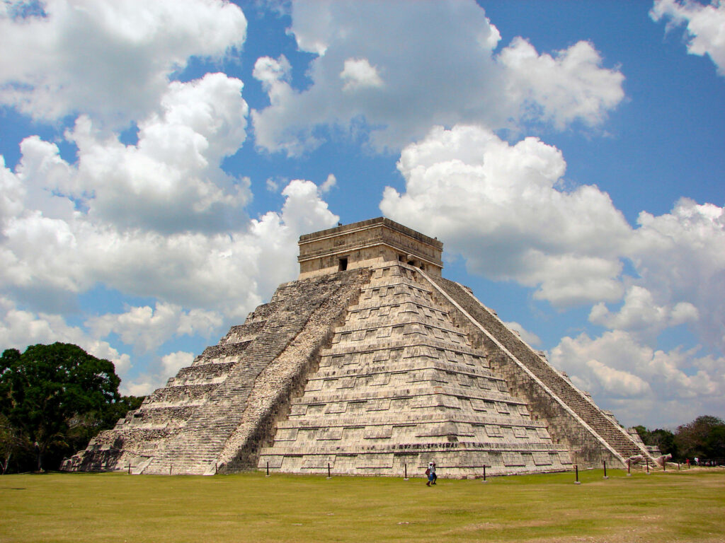 Chichen Itza pyramid in sunlight with blue sky white clouds and lush green grass background Wallpaper