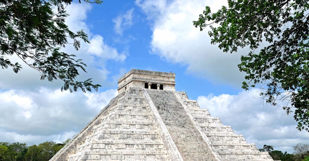 Chichén Itzá pyramid framed by green trees under blue sky with clouds ancient stone architecture Wallpaper