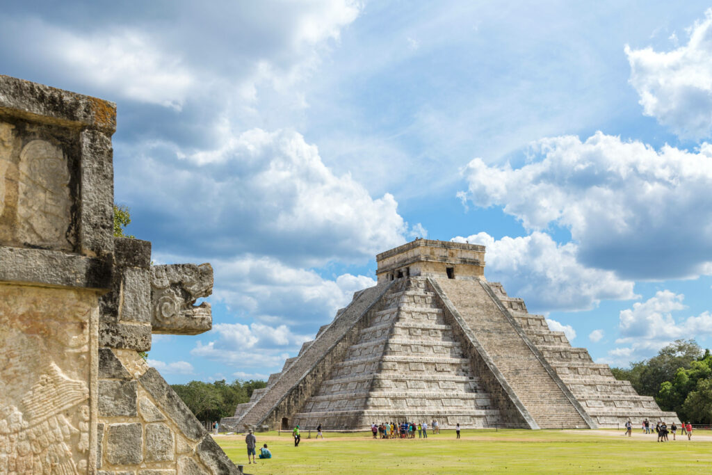 Tourists exploring Chichen Itza with stepped pyramid stone carvings lush greenery blue sky background Wallpaper