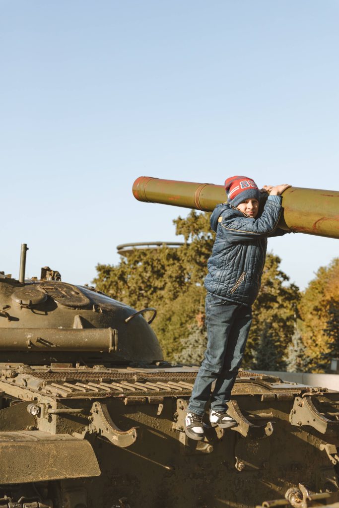 Young boy in winter hat playfully holds weathered military tank barrel against blue sky background Wallpaper