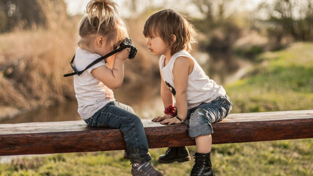 Two children on wooden beam playing with camera outdoors in warm sunlight natural setting wallpaper