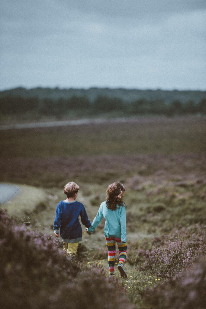Two children holding hands walking in colorful clothes through open field with rolling hills Wallpaper
