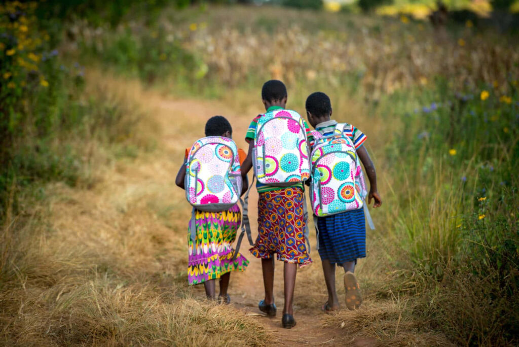 Three children with colorful backpacks walk hand in hand on a sunlit wildflower path outdoors Wallpaper