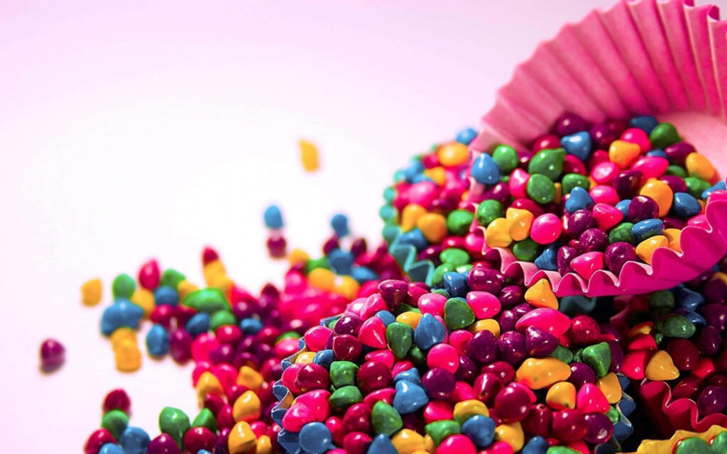 Colorful choco pebble candy spilling from pink cupcake liner on pastel dessert-themed wallpaper