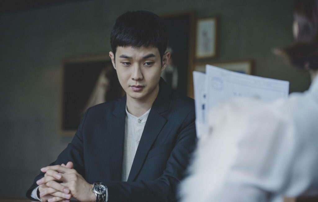 Actor Choi Woo-shik in dark suit and watch seated thoughtfully during intense conversation wallpaper background
