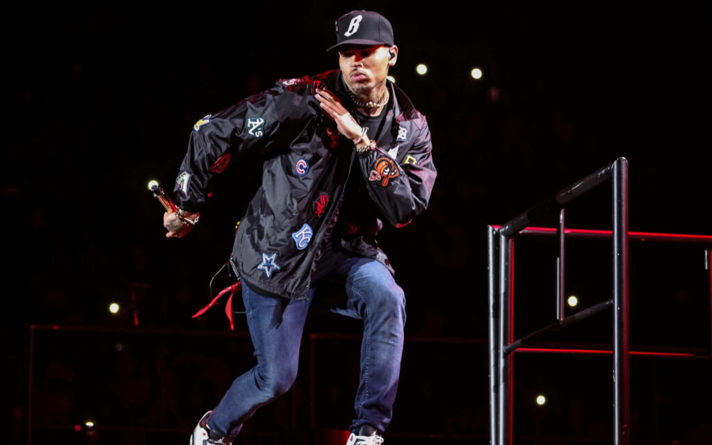 Chris Brown energetic on stage in stylish jacket and cap captivating audience wallpaper background