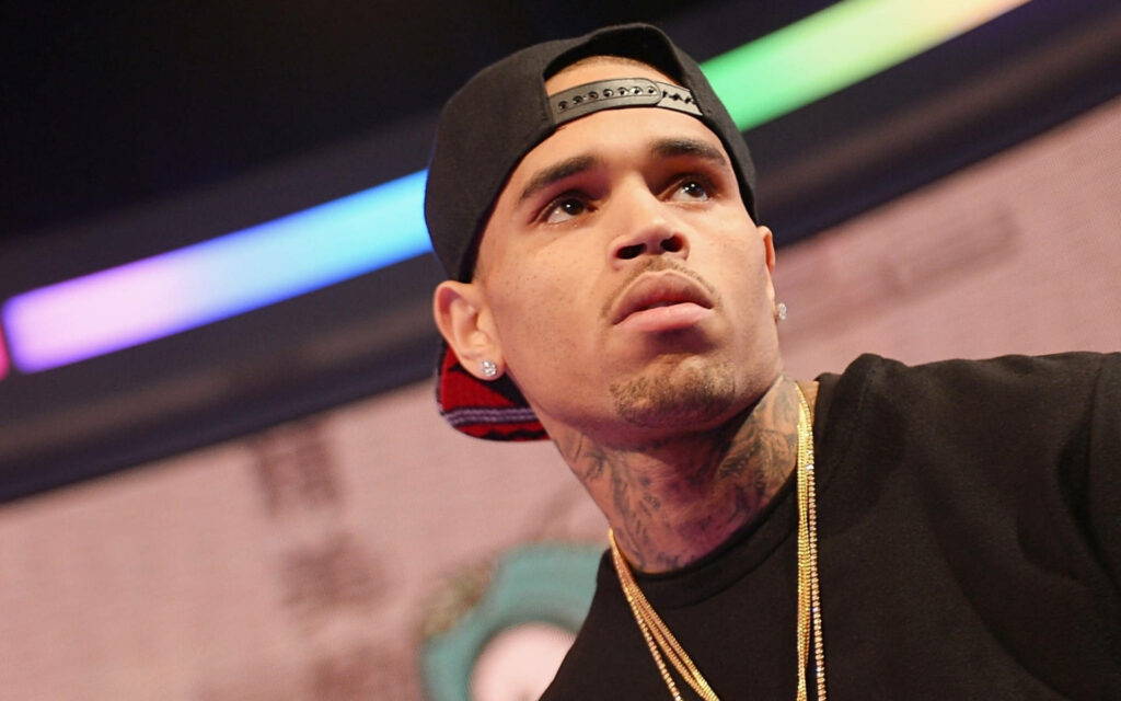 Low angle photo of Chris Brown in black cap gold chains with vibrant colorful lights background Wallpaper