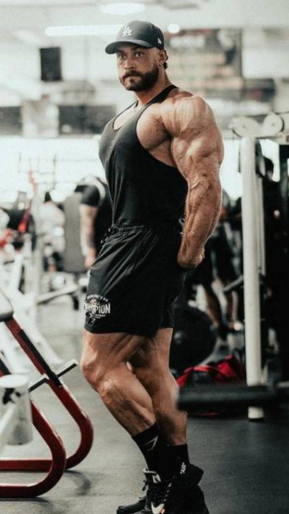 Chris Bumstead side profile gym pose showcasing muscular physique black tank cap wallpaper background