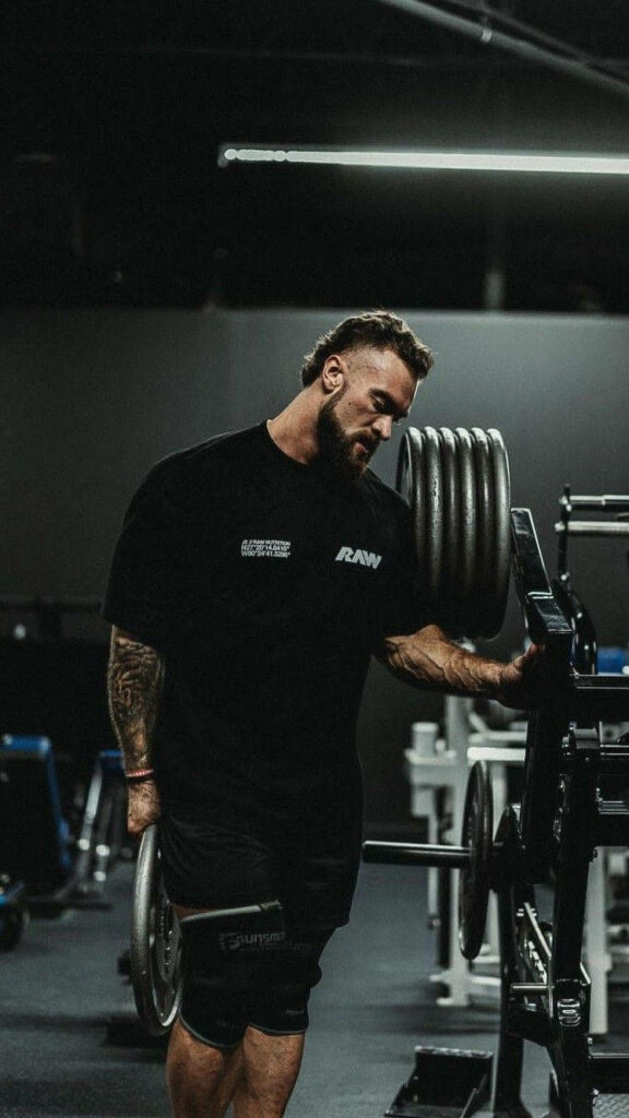 Chris Bumstead in dimly lit gym holding weight plate showcasing muscular physique and tattoos Wallpaper