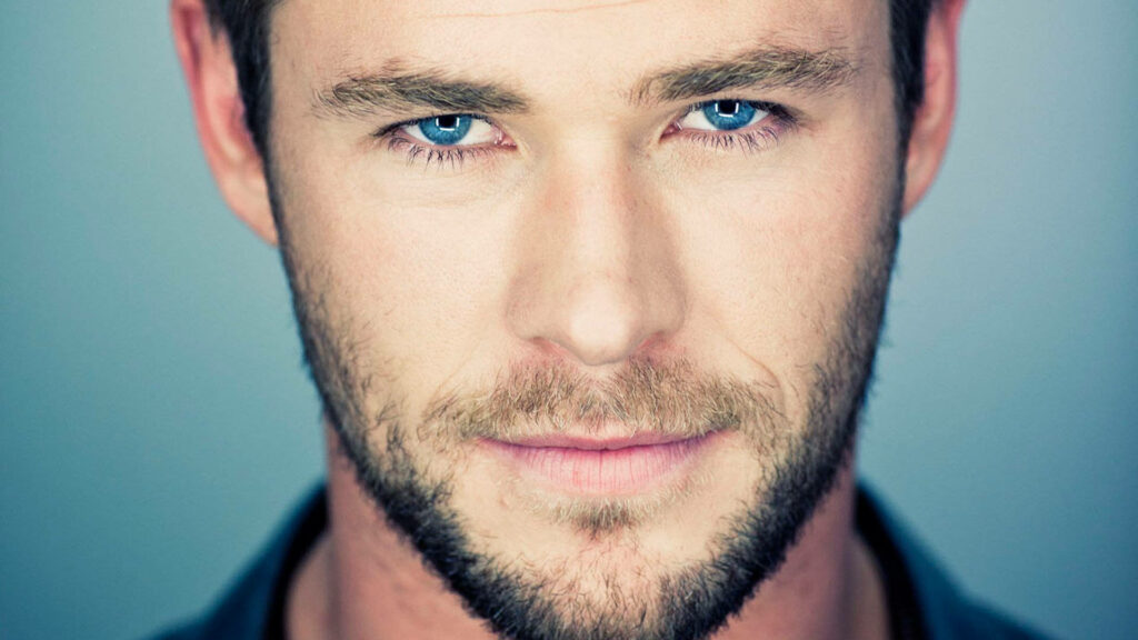 Close-up of Chris Hemsworth smirking with striking blue eyes and rugged beard wallpaper background