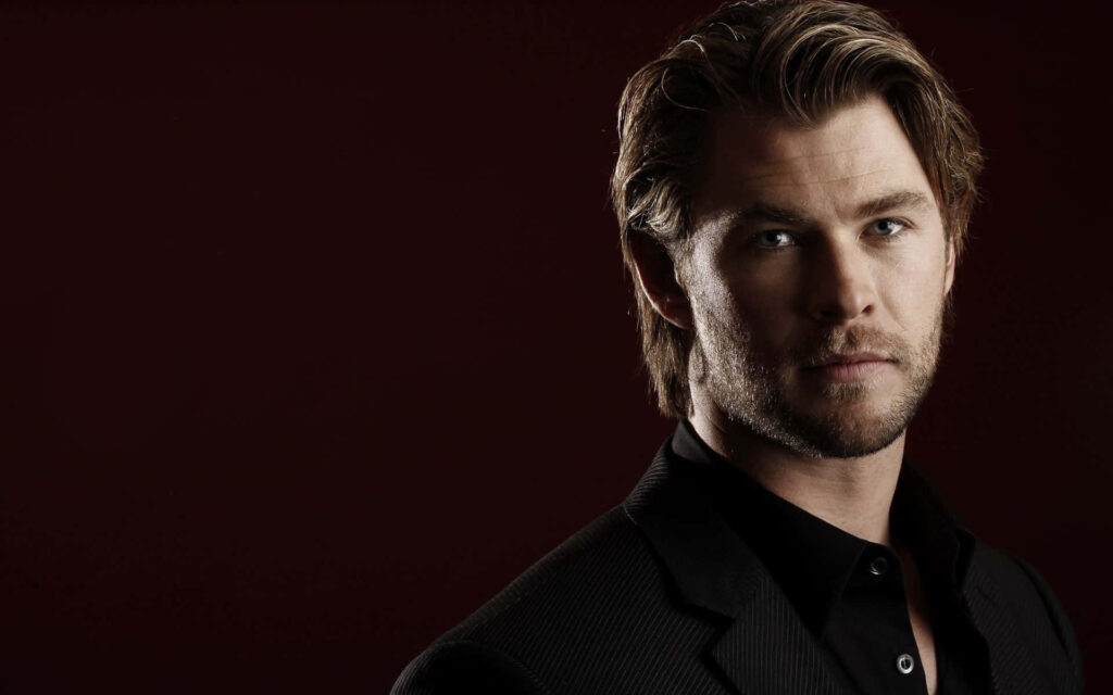 Chris Hemsworth in dark suit posing confidently against deep maroon background wallpaper