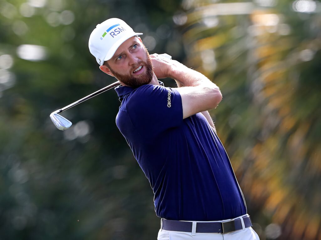 Chris Kirk mid-swing golf stance navy shirt white cap sunlit lush golf course background wallpaper